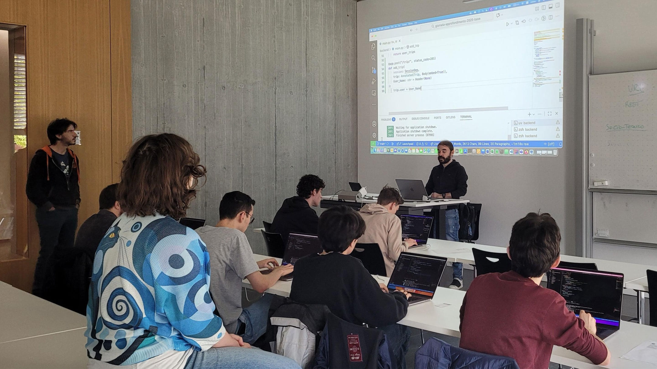 Students of Liceo di Mendrisio coding on their laptops in a classroom at the USI Faculty of Informatics, while an instructor live-codes a Python back end projected on the wall