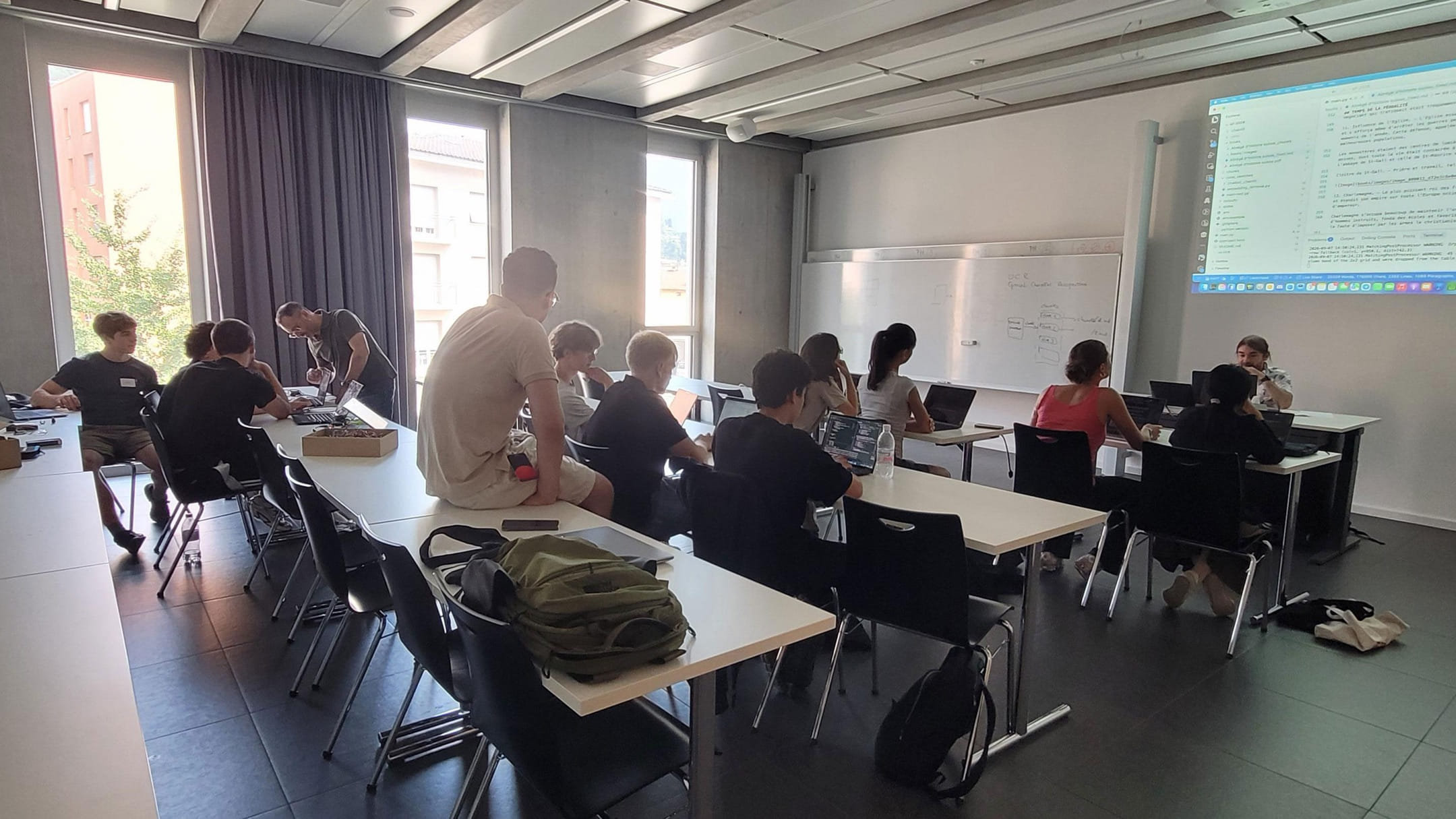 Coding Camp participants working on their laptops in a classroom at the USI Faculty of Informatics, with a code editor projected on the wall
